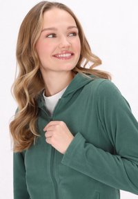 Felpa verde in pile con zip frontale, cappuccio e maniche lunghe. Texture morbida, design casual e silhouette aderente.