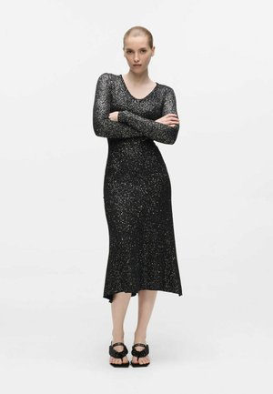 Femme aux cheveux tirés en arrière, portant une robe midi noire pailletée à manches longues et des sandales noires, debout les bras croisés devant un fond blanc.