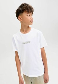 Witte katoenen t-shirt met korte mouwen met een klein, groen geborduurd logo op de borst. Het model draagt een lichtbeige broek.