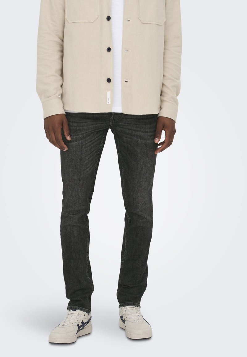 Camicia beige a bottoni sopra una maglietta bianca, abbinata a jeans slim-fit grigio scuro e sneakers bianche con dettagli blu.
