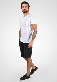 Weißes T-Shirt mit schwarzen Punkten, kombiniert mit schwarzen Shorts und schwarzen Sneakern. Das Outfit hat ein lässiges Design und einen schmalen Schnitt.