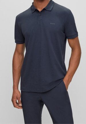 Mann in einem dunkelblauen Poloshirt mit kurzen Ärmeln und dem "BOSS"-Logo auf der linken Brust, passende dunkelblaue Hose, Hand in der Tasche.