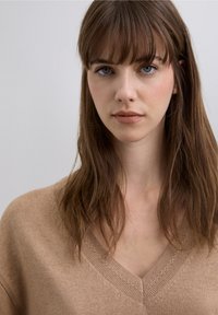 Suéter de cuello en V color marrón claro con cuello acanalado y sutiles detalles texturizados. La tela suave cae delicadamente alrededor de los hombros y la clavícula.