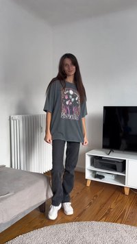 Hallikas suuruses T-särk, millel on lilleline graafika ja tekst, koos mustade sirgete pükste ja valgete tossudega puidust põranda peal.