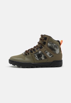 Bottes de randonnée montantes en vert olive avec semelle en caoutchouc noire, accents en maille camouflage, lacets réglables orange et noir, et œillets en métal.