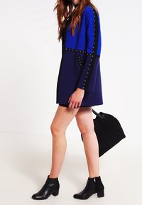 Kort klänning i blått och marinblått med nitar längs ärmarna och fickorna. Bärs med svarta ankelboots och en svart handväska.