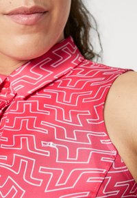 Hihaton poolopaita kirkkaassa vaaleanpunaisessa, jossa on valkoinen geometrinen kuvio. Sisältää kauluksen, nappisulkemisen ja pienen logon kankaassa.