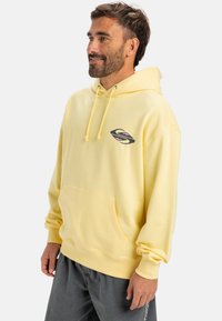 Yellow hooded sweatshirt made of soft fabric, featuring a front pocket and a stylized logo in black and pink on the chest.