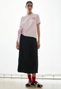 Rosa Baumwoll-T-Shirt mit rotem Schriftzug, kombiniert mit einem dunkelblauen Jeansrock. Schwarze Sneaker mit roten Socken. Einfaches Design, lässige Passform und entspannter Stil.