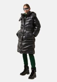 Black puffer coat with a waist cinch and hood, featuring a glossy texture. Paired with green pants and black fur-lined boots.
