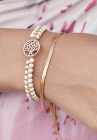 Two wristbands on a wrist: one with white beads and a circular metal charm featuring a tree design; the other is a smooth, copper-colored band.