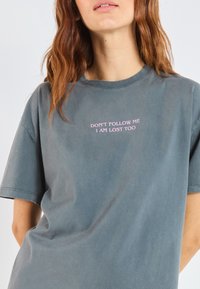 Žalias medvilninis marškinėliai trumpomis rankovėmis ir apvalia iškirpte. Priekyje, krūtinėje, yra rožinės spalvos tekstas "NENUEIK PAS MANE, AŠ IRGI PASIKLYDĘS".