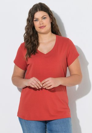 Frau mit langen lockigen Haaren, die ein rotes T-Shirt mit V-Ausschnitt und blaue Jeans trägt, steht mit verschränkten Händen an einer weißen Wand.