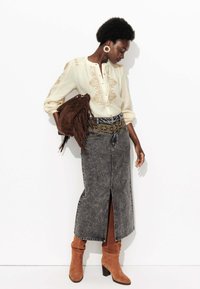 Jupe longue en denim gris avec une fente à l'avant, associée à un chemisier crème avec broderies dorées. Bottes à talons en daim marron et sac à franges.