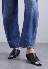 Mule in pelle nera con punta affilata, dotate di un accento di borchie argentate sulla punta. Abbinati a jeans in denim a gamba larga e dal lavaggio chiaro.