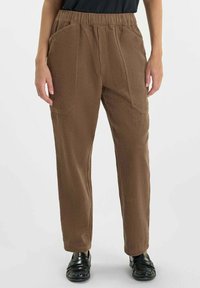 Persona vistiendo pantalones rectos marrones texturizados con bolsillos laterales y zapatos negros sin cordones, de pie sobre un fondo sencillo.
