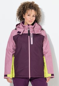 Frau mit lockigem Haar trägt eine lila, pinke und gelbe Winterjacke mit hohem Kragen und Reißverschlusstasche auf der Brust, steht vor einer weißen Wand.