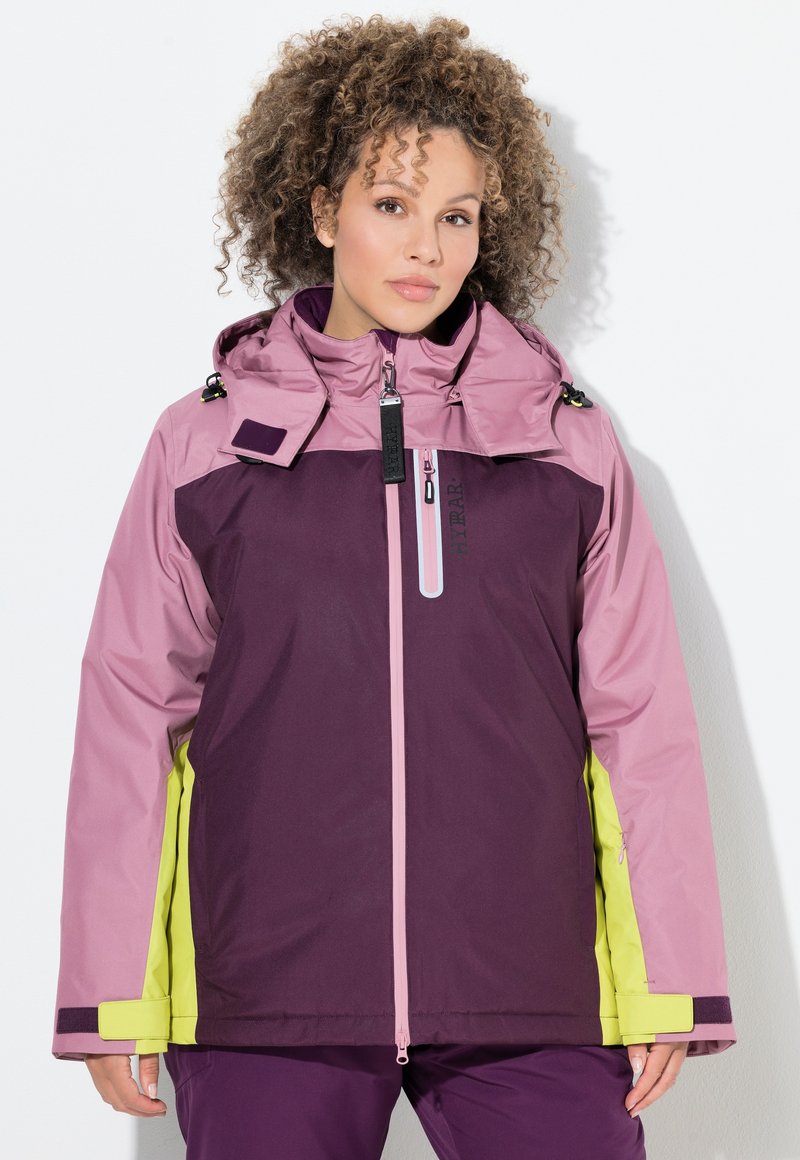 Frau mit lockigem Haar trägt eine lila, pinke und gelbe Winterjacke mit hohem Kragen und Reißverschlusstasche auf der Brust, steht vor einer weißen Wand.