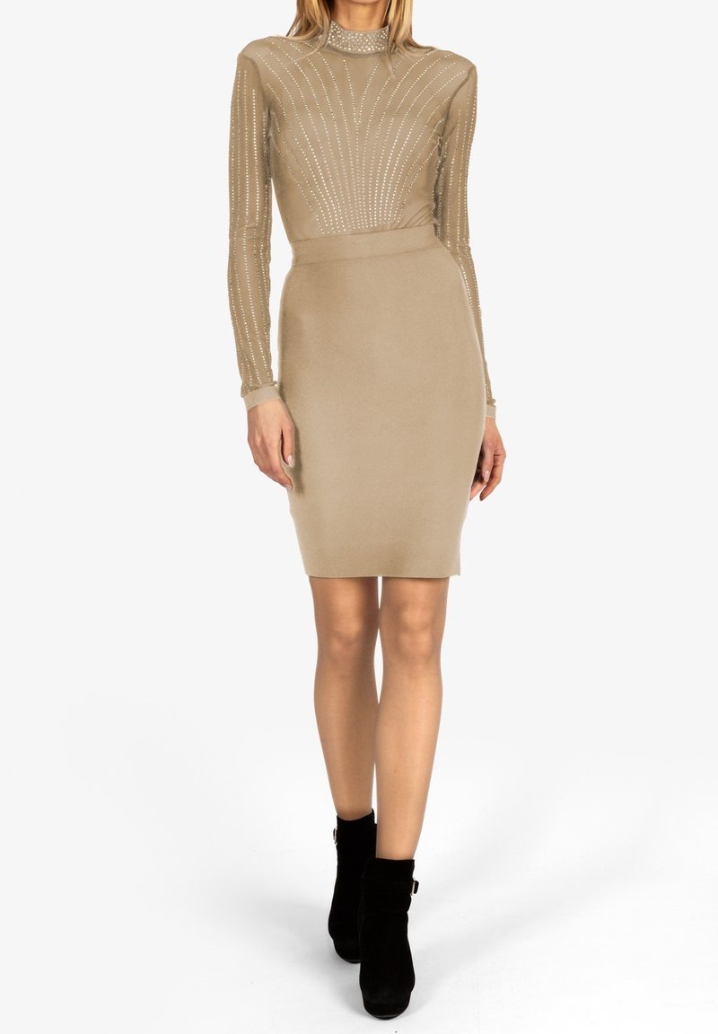 Femme portant une robe beige ajustée avec un haut transparent à motifs et une jupe unie, associée à des bottines noires, debout sur un fond blanc.