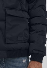 Svart polstret jakke med en frontlomme, ribbede mansjetter og teksturert stoff. Inkluderer en merkelapp på lommen. Brukt over slitte jeans.