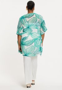 Durchsichtiger Kimono mit grünem und tealfarbenem Blumenmuster, mit breiten Ärmeln und seitlichen Schlitzen, kombiniert mit weißen, geraden Hosen und Absätzen.
