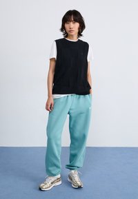 Haut sans manches noir texturé sur un T-shirt blanc, associé à un pantalon de jogging teal et des chaussures de course à accents dorés. Design simple, look décontracté.
