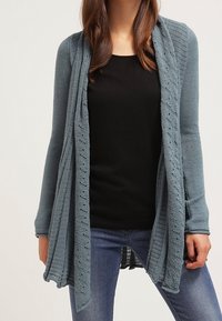 Långärmad cardigan i mjuk blå stickning med öppen front, ribbade detaljer och spetsdekoration längs fållen och ärmarna. Bärs över en svart topp.