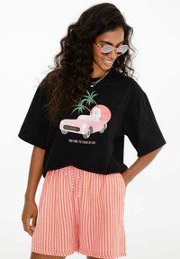 Žena s dlouhými kudrnatými vlasy, nosící kulaté sluneční brýle, černé tričko s grafikou růžového auta a textem, a šortky s oranžovými pruhy a šňůrkou.