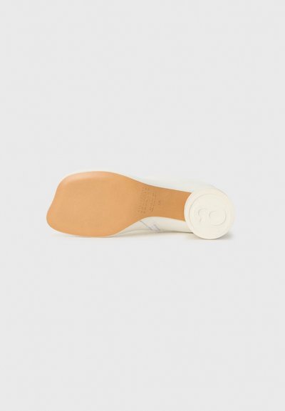 Chaussure couleur crème avec une semelle en caoutchouc, présentant une texture lisse et un talon arrondi. Le dessous montre un logo en relief et des indications de taille.