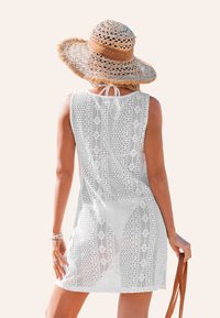 Robe de couverture en crochet blanche avec un design ample sans manches, ornée de motifs floraux et d'un lien au cou. Portée avec un chapeau en paille.
