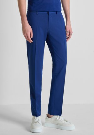 Hombre con pantalones ajustados azul real y zapatillas blancas de pie contra un fondo claro y simple.