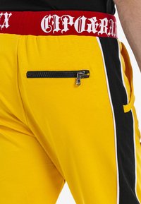 Gelbe Jogginghosen mit einer schwarzen Reißverschlusstasche, einem roten Bund mit weißem Text und schwarzen Seitenpaneelen mit weißen Streifen.