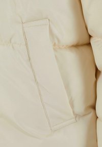 Béžová puffer bunda z látky s textúrovaným povrchom, s nášivkovou vreckom a viditeľným prešívaním pre trvácnosť a štrukturálne detaily.