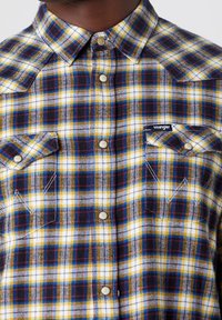 Geruite flanellen overhemd in gele, blauwe en rode tinten. Bevat een puntige kraag, drukknoopsluitingen en twee borstzakken met kleppen.