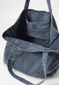 Blå denim tote bag med doble håndtak, en innerlomme merket "GAP" og en avslappet, uformet form.