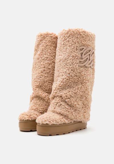 Pitkät, pörröiset beige saappaat, jotka on valmistettu teksturoidusta lampaanvillasta, paksulla ruskealla alustalla ja koristeellisella logolla sivussa.
