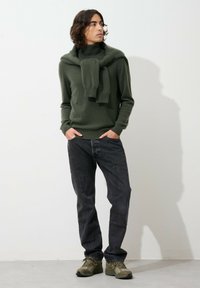 Maglione verde lavorato a maglia con collo alto e una sciarpa drappata sulle spalle, abbinato a jeans scuri e sneakers verdi, su uno sfondo neutro.