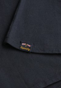 Marineblåt bomulds T-shirt-stof med et synligt Superdry-mærke, der har britiske og amerikanske flag i gule og blå sy-detaljer.