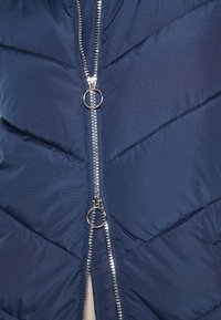 Ναυτικό μπλε γιλέκο τύπου puffer με σχεδίαση σε μορφή βέλους, διαθέτει ασημένιο φερμουάρ, με κρίκο για εύκολη χρήση και απλή, ματ υφή.