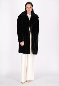 Sort faux fur-frakke med bred krave og lange ærmer. Modellen har en hvid bluse og lyse bukser på og står foran en ensfarvet baggrund.