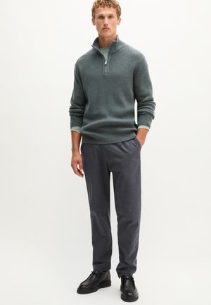 Mann steht und trägt einen grauen, gerippten Viertel-Zip-Pullover, dunkelgraue Hose und schwarze Schuhe vor einfachem hellem Hintergrund.