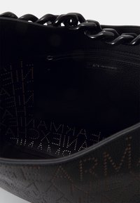 Svart tote-väska med en strukturerad yta, som har en stor innerficka och ett mönster av präglad branding. Handtag av kraftiga kedjelänkar.