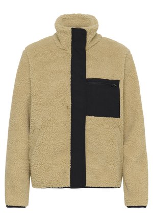 Veste en polaire sherpa beige avec col haut, couverture de fermeture éclair noire à l'avant et poche poitrine zippée noire.