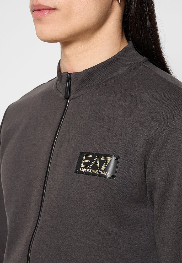 Felpa grigia con zip e colletto alto. Presenta un badge del logo EA7 Emporio Armani nero e oro sul lato sinistro del petto. Tessuto morbido e testurizzato.