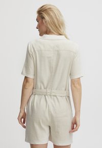 Combinaison en lin courte de couleur beige clair. Elle présente des manches courtes, une taille ceinturée et un col pointu. Texture douce avec poches arrière.
