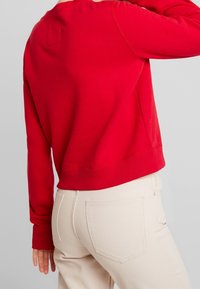 Sudadera roja con cuello redondo y mangas largas, que presenta puños y dobladillo acanalados. Llevada sobre pantalones beige claros con detalle en el bolsillo trasero.