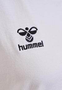 Logo de la marque hummel brodé en noir avec une abeille stylisée sur tissu blanc, centré et bien visible.