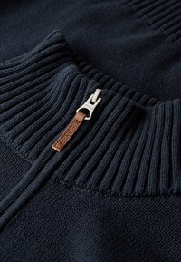 Navy Strickpullover mit geripptem Kragen, Reißverschluss mit silberner Hardware und einem braunen Lederakzent-Tag. Weiche Textur im gesamten Pullover.
