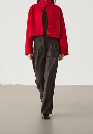 Osoba idąca ubrana w krótki czerwony zapinany na guziki żakiet, ciemne plisowane spodnie z wysokim stanem oraz czarne buty na jednolitym szarym tle.