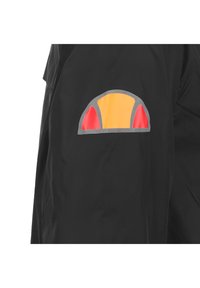 Zwarte jas met een kleurrijke patch op de mouw, voorzien van een ontwerp in geel, rood en grijs. Het materiaal lijkt lichtgewicht en soepel.